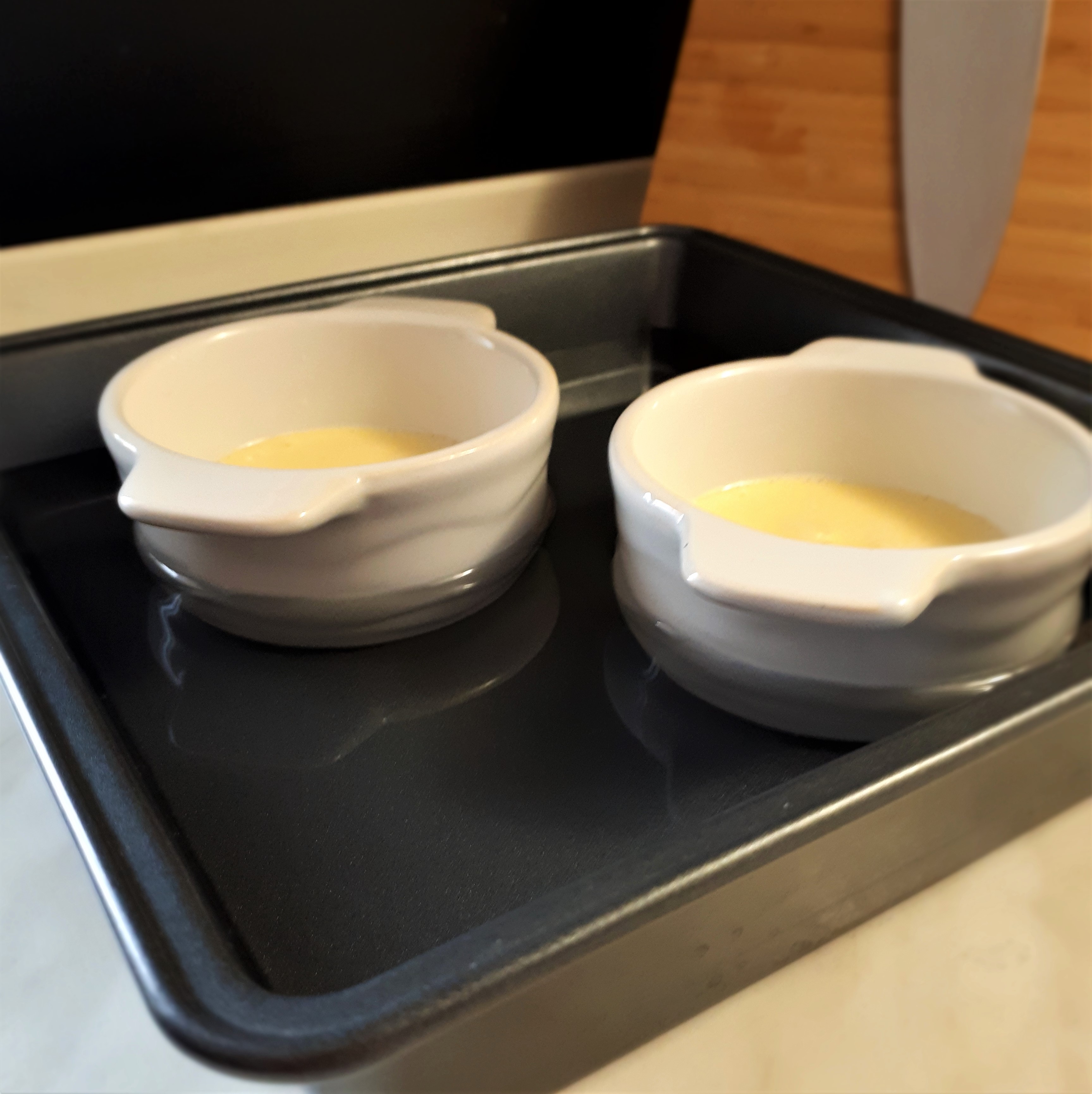 Dve porcie Crème brûlée v bielych zapekacích miskách vo štvorcovej forme na pečenie zaliate vodou do polovice misiek.