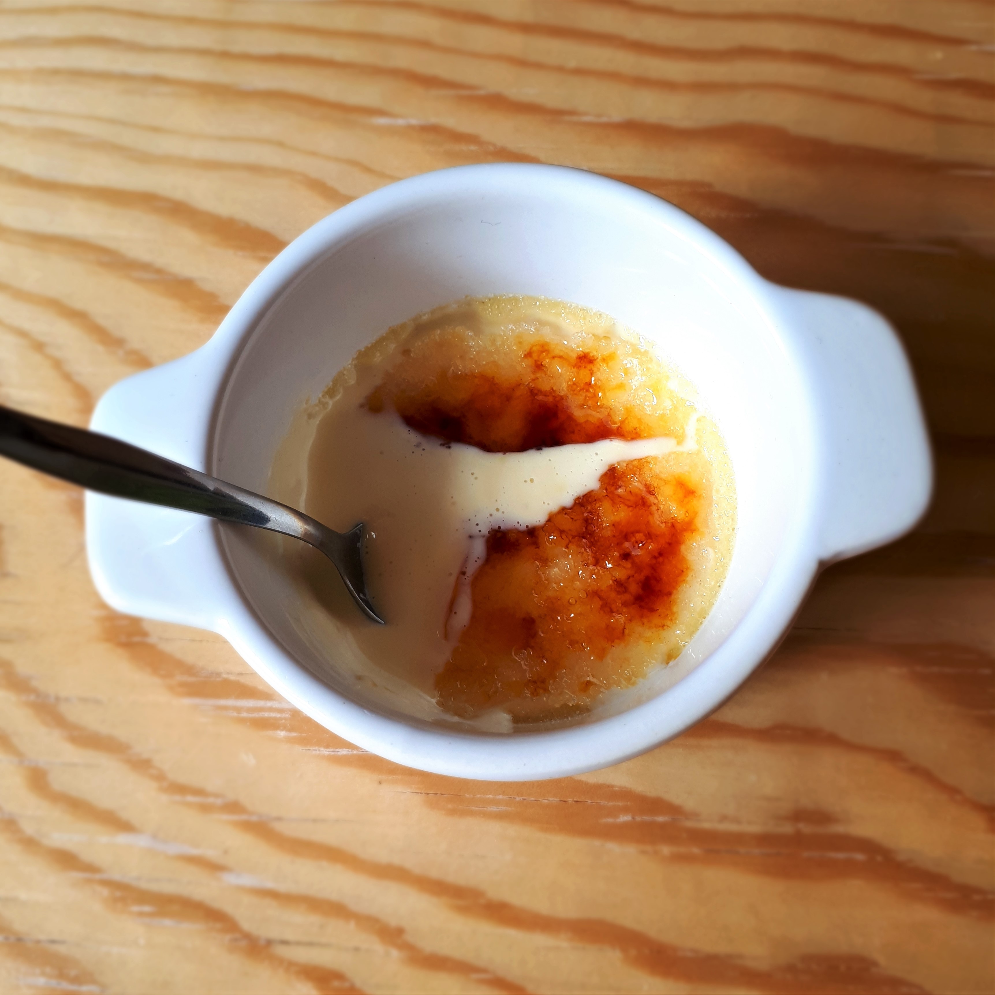 Jedna porcia Crème brûlée na drevenom stole karamelová kôrka na vrchu Crème brûlée rozbitá lyžičkou namočenou v svetlom kréme.