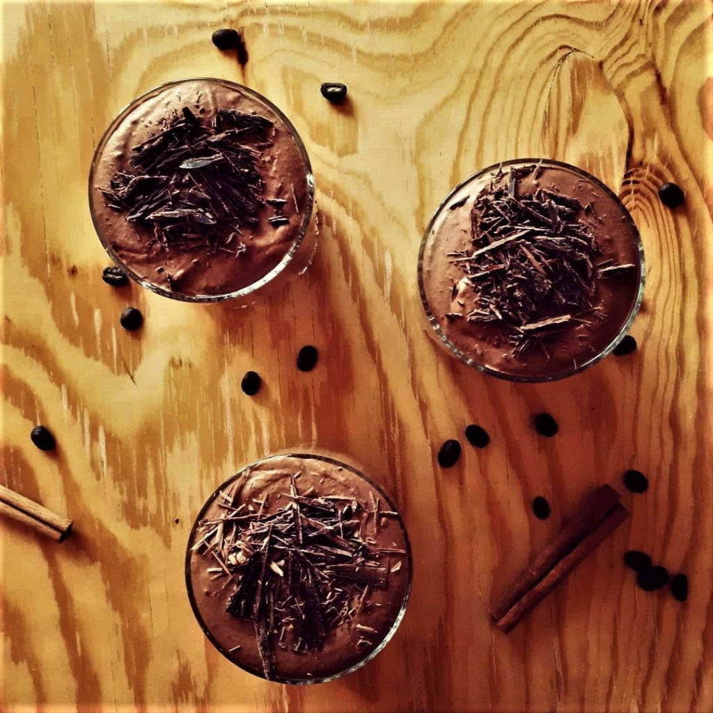 3 poháre gaštanovo-čokoládovej peny na drevenom stole dozdobené čokoládovými hoblinkami a obsypané kávovými zrnami a škoricou.