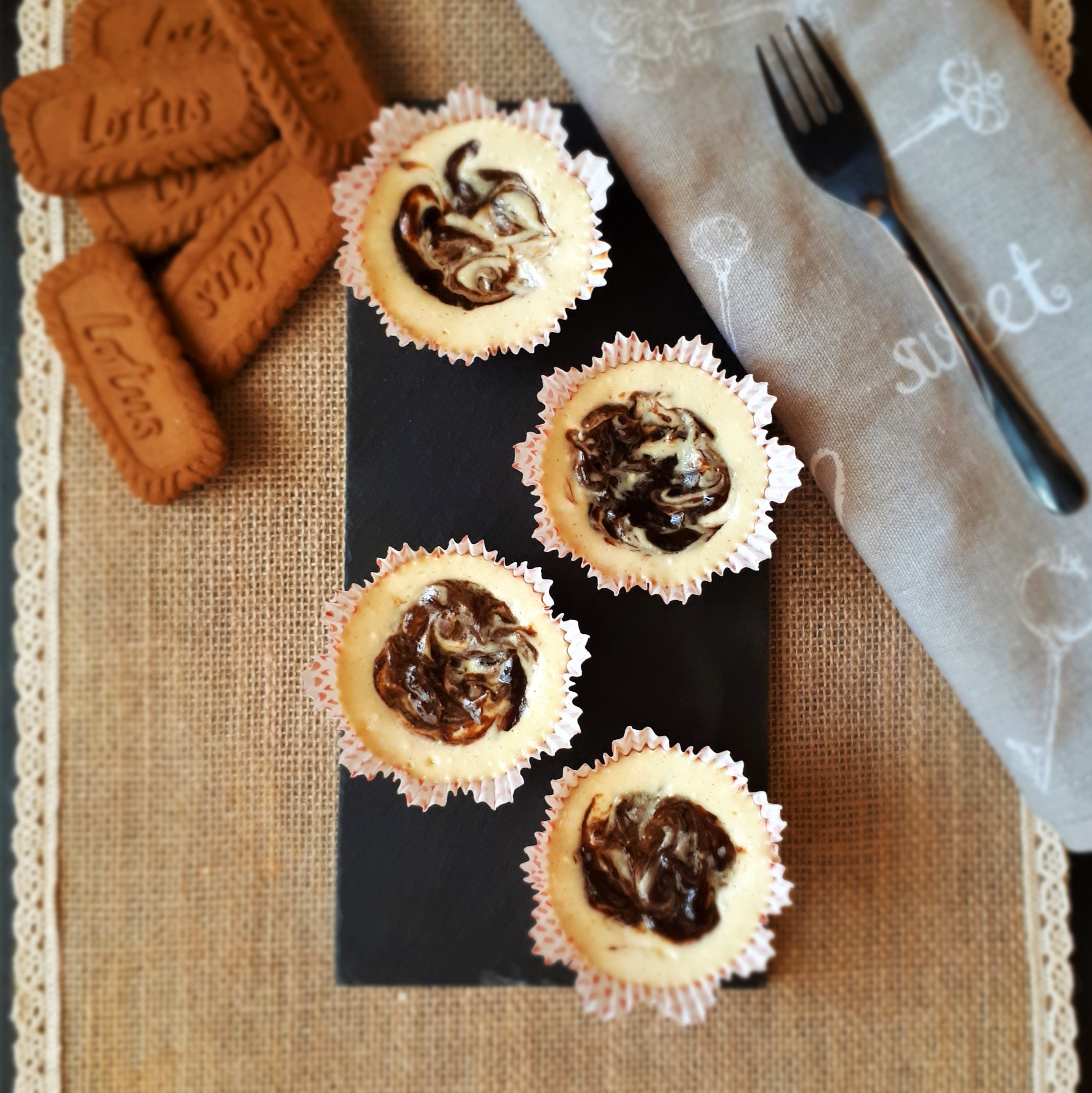 Štyri mini cheesecake muffiny so slivkovým lekvárom v bielych papierovým košíčkoch na čiernej kamennej doske plstenné prestieranie šedá kuchynská utierka s dezertovu lyžičkou vysypané Lotus sušienky