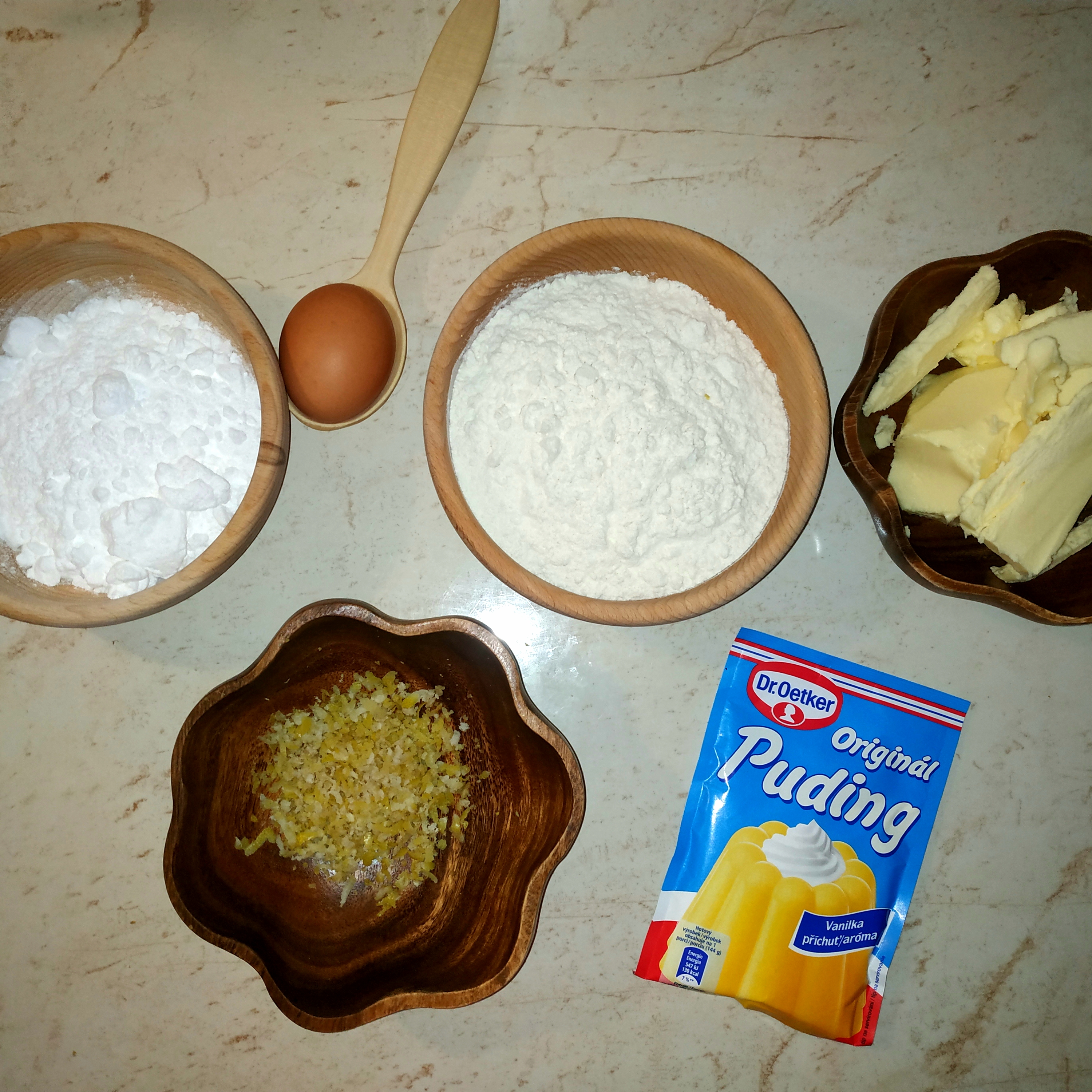 Drevená miska s múkou drevená miska s cukrom drevená lyžica s vajíčkom drevená miska s maslom a nastrúhanou citrónovou kôrou vanilkový puding na mramorovom povrchu