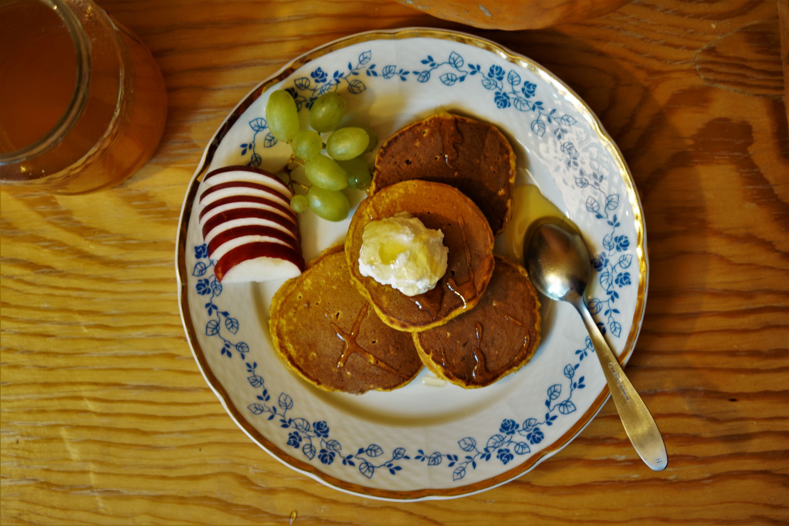 Štyri oranžové lievance navrchu kopček tvarohu pokvapkané medom na bielom tanieri s modrými kvetinami a zlatým okrajom nakrájané jablko a strapec hrozna v pozadí tekvica a pohár s medom na drevenom stole