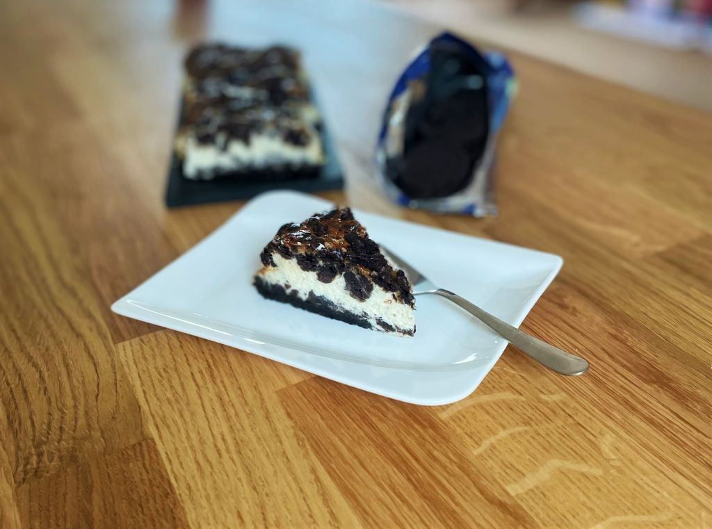 Kúsok Oreo cheesecake na bielom dezertnom tanieri dezertná vidlička v pozadí balíček Oreo sušienok a na čiernej doske zvyšné kúsky torty na drevenom stole
