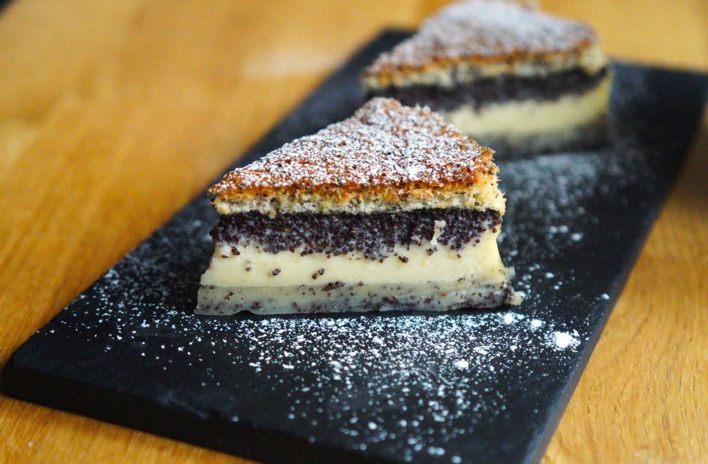 Trojvrstvý makový koláč posypaný práškovým cukrom na čiernom tanieri a drevenom stole v pozadí ďalší kúsok koláča